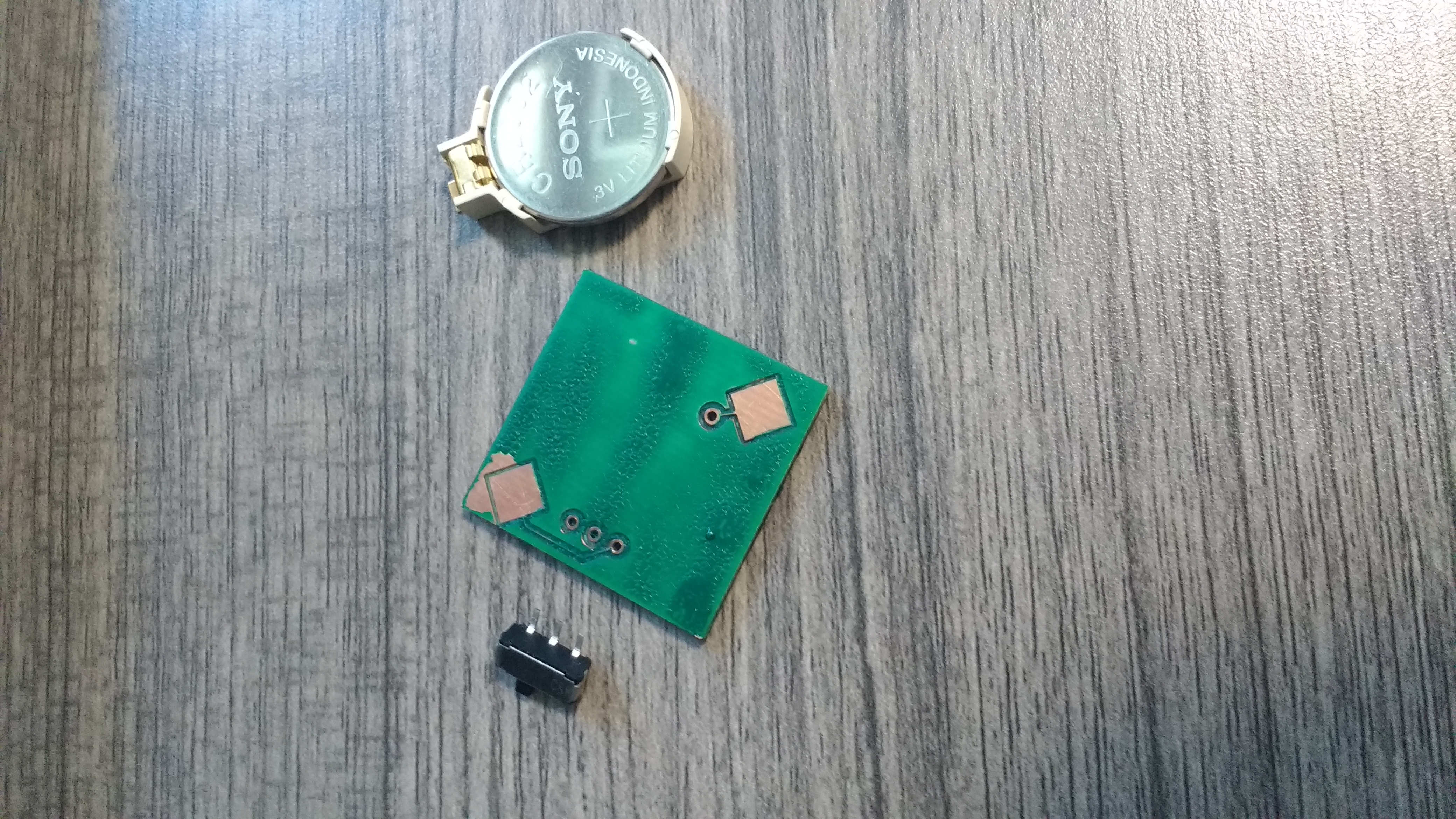the bottom of a 1 inch by 1 inch copper PCB. It has green insulation on it, with large exposed copper pads for soldering a battery mount.